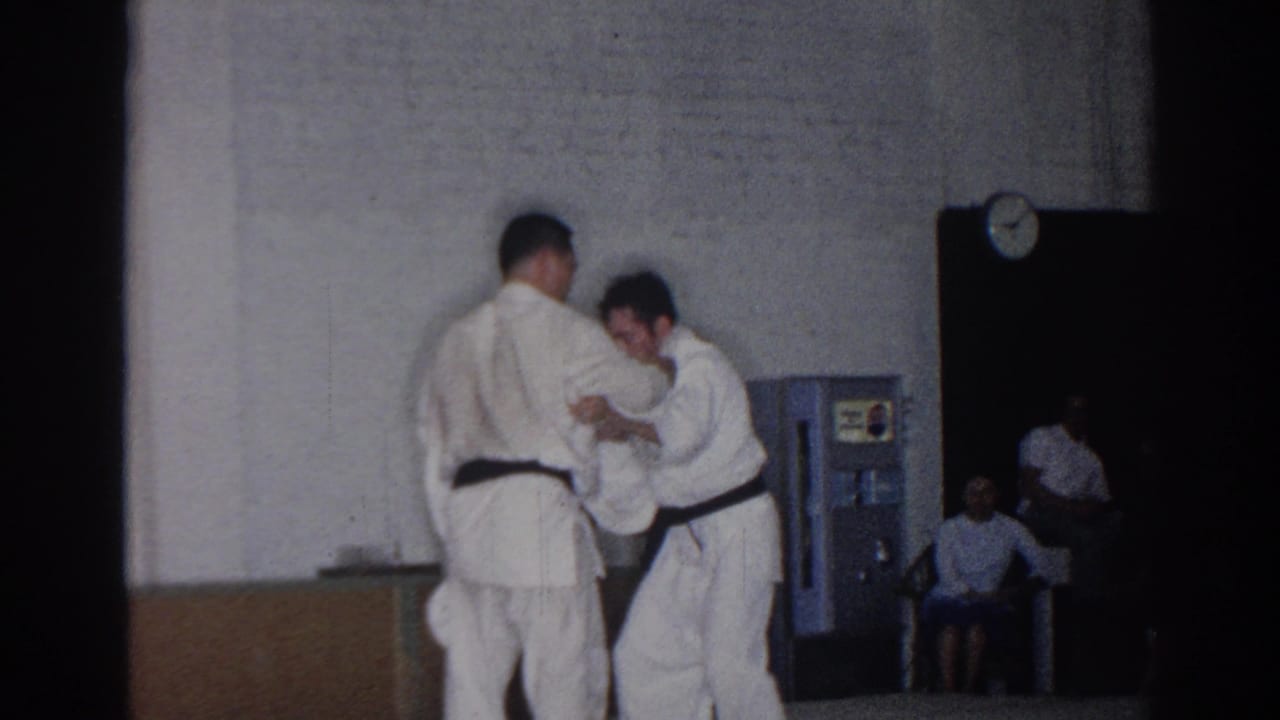 Two men in white gi and black belts stand close together in a gym while spectators sit near the wall.