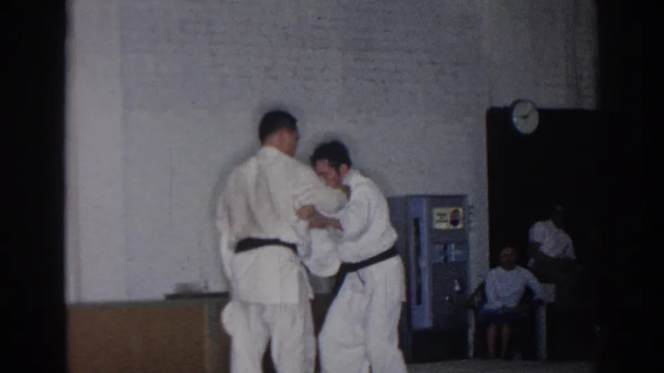 In 1966 San Gabriel, two martial artists in white gi and black belts begin a judo exchange inside a gymnasium with seated spectators nearby.
