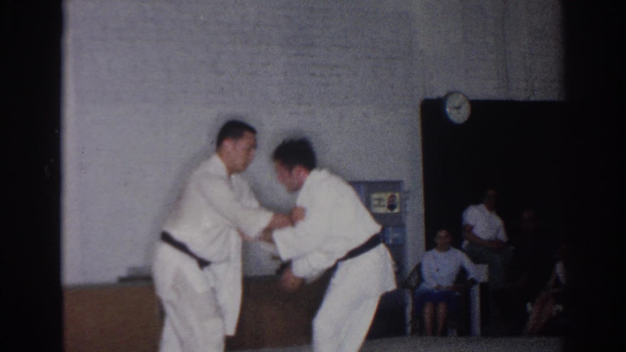 Two men in white gi practice a judo technique in a San Gabriel gymnasium, with chairs, a wall clock, and observers visible behind them.