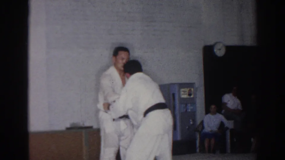 A judo practitioner bends into a throw attempt while his partner remains upright inside the 1966 San Gabriel gym setting.