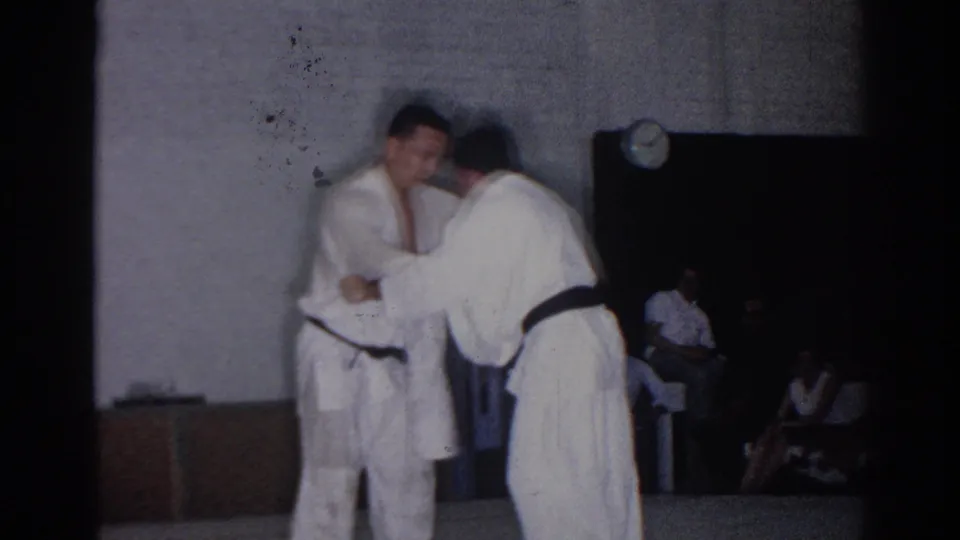The judo pair continues close contact in the gymnasium as spectators sit near a dark doorway and a wall clock remains visible.