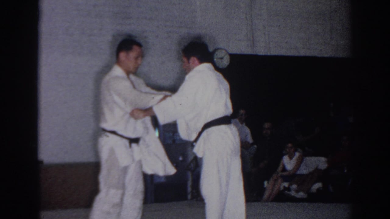 Two men in white karate gi grip each other in an indoor gym with spectators behind them.