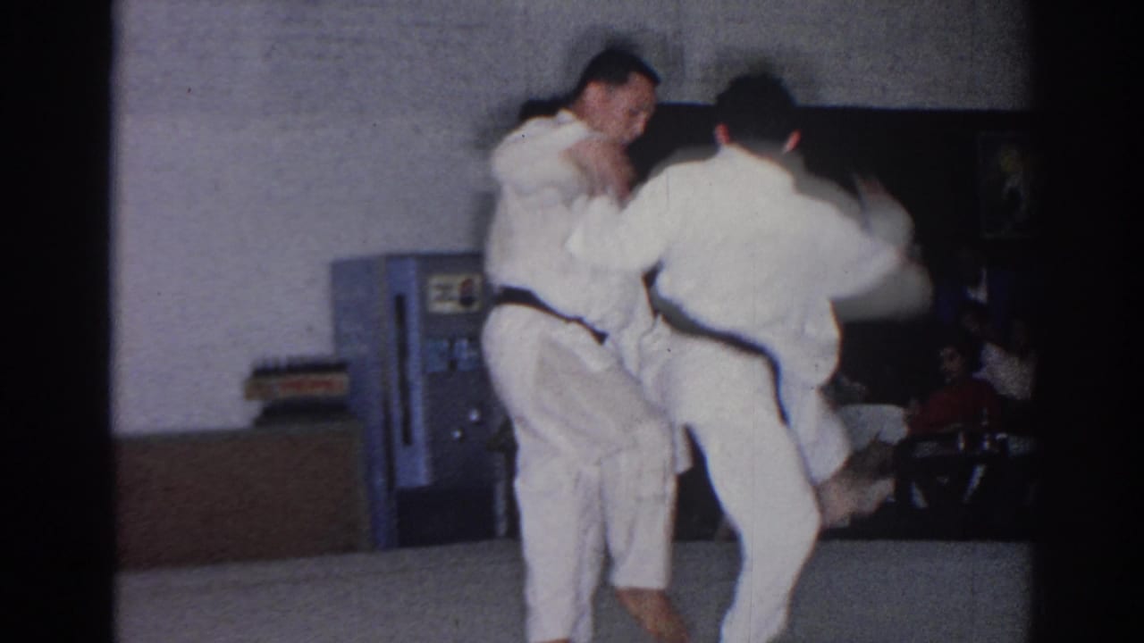 Karate competitors move through a throw or lift in front of a gym wall and seated audience.