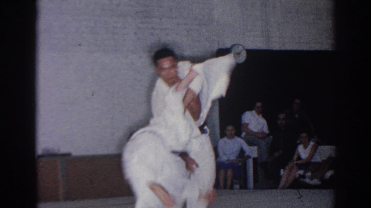Two karate fighters in white gi grapple on a mat as spectators sit behind them.