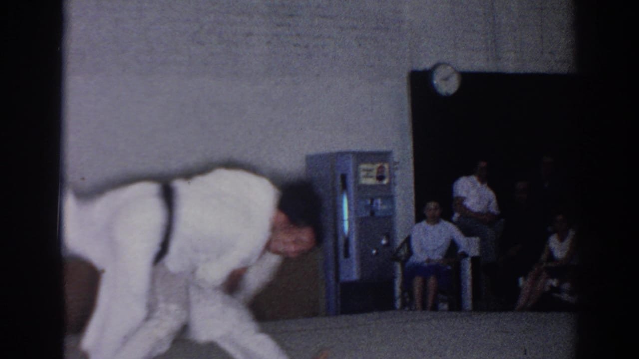 A karate fighter bends forward on the mat with spectators, a vending machine, and a wall clock behind him.