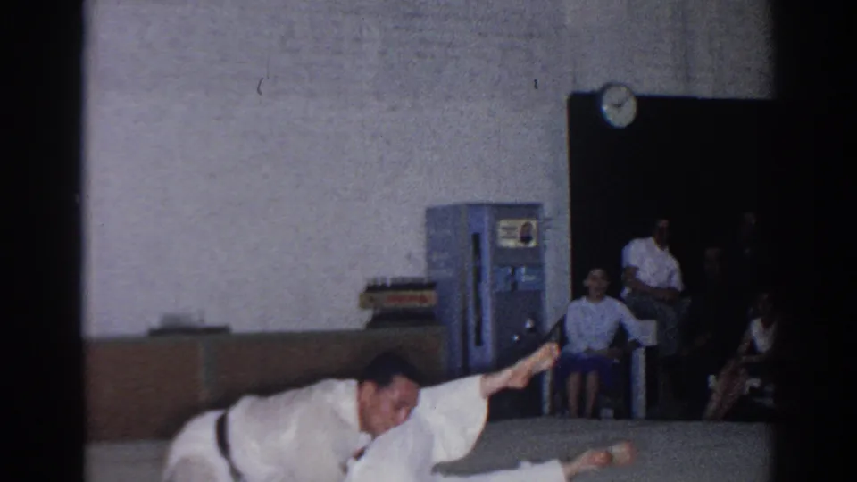 During the 1966 San Gabriel karate bout, a fighter in a white gi crouches and extends one arm as spectators sit along the wall.