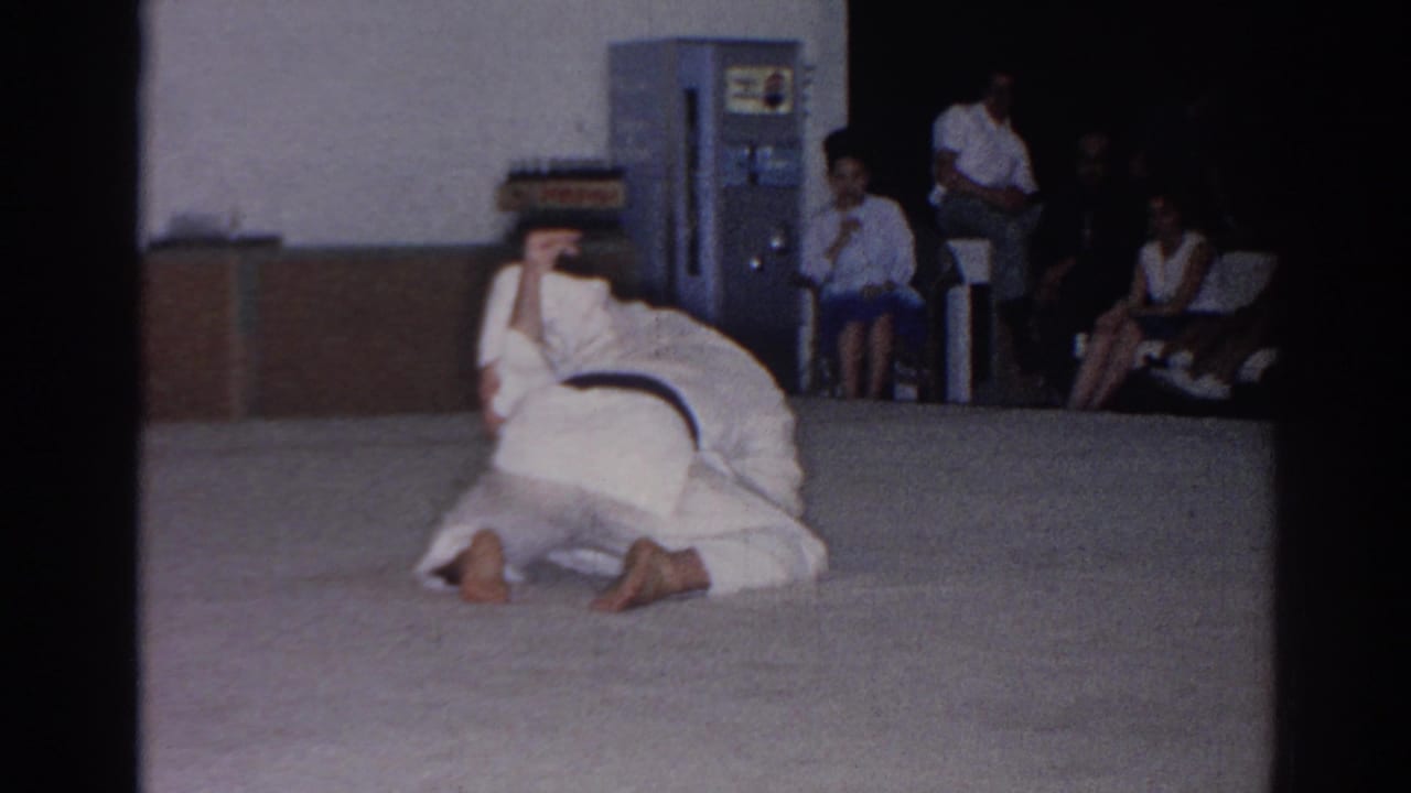 In San Gabriel, California, 1966, two martial artists in white gi demonstrate a hold or throw on a mat as spectators watch indoors.