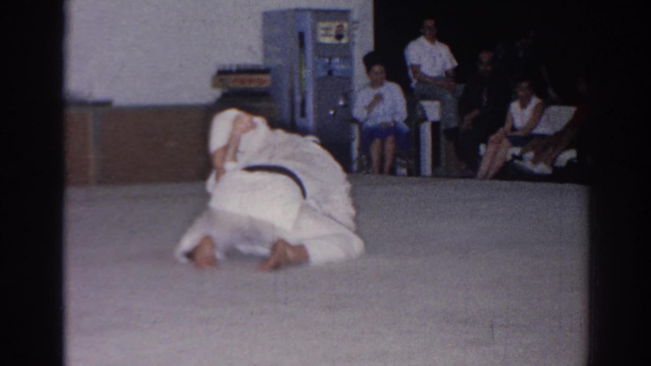 A 1966 San Gabriel martial-arts demonstration shows one participant kneeling over another on a mat, with an audience seated near a vending machine.
