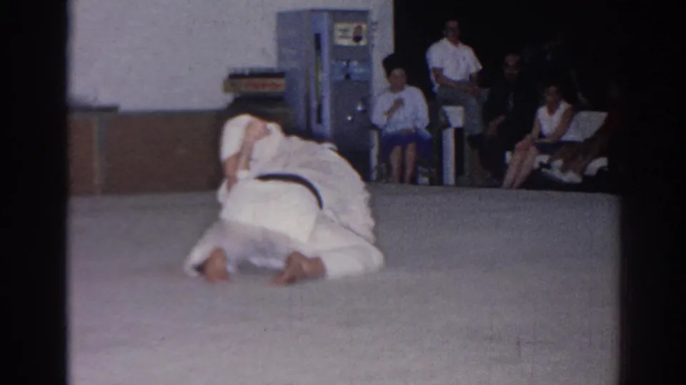 A 1966 San Gabriel martial-arts demonstration shows one participant kneeling over another on a mat, with an audience seated near a vending machine.