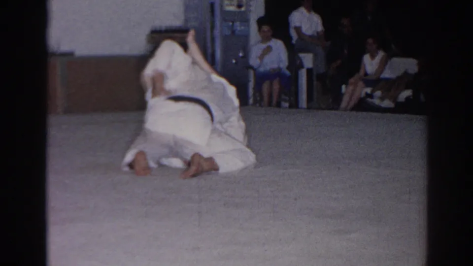 The 8mm frame shows a close view of two white-clad martial artists on a mat in San Gabriel, with seated spectators along the back wall.