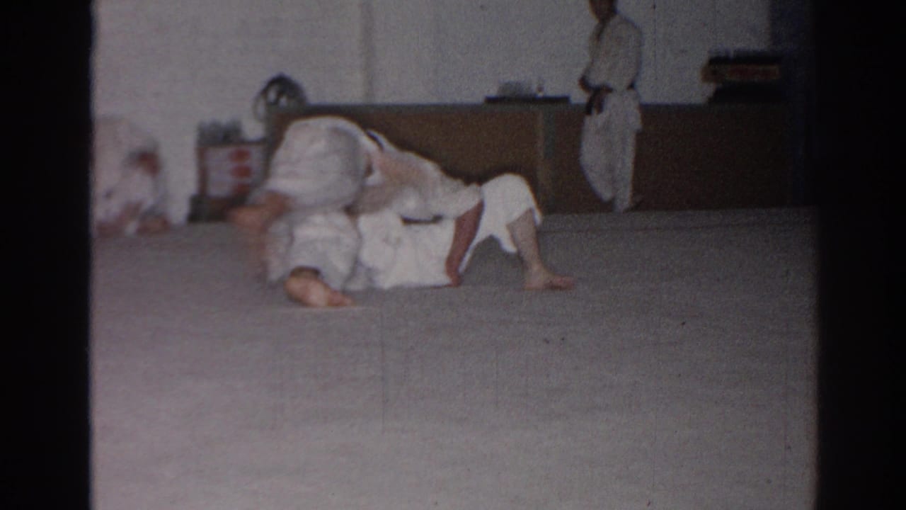 Two people in white gis grapple on a mat while another participant stands nearby.