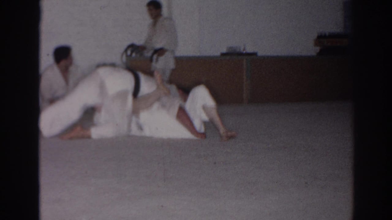 A martial artist in a white gi moves over a grounded partner as others watch from the side.