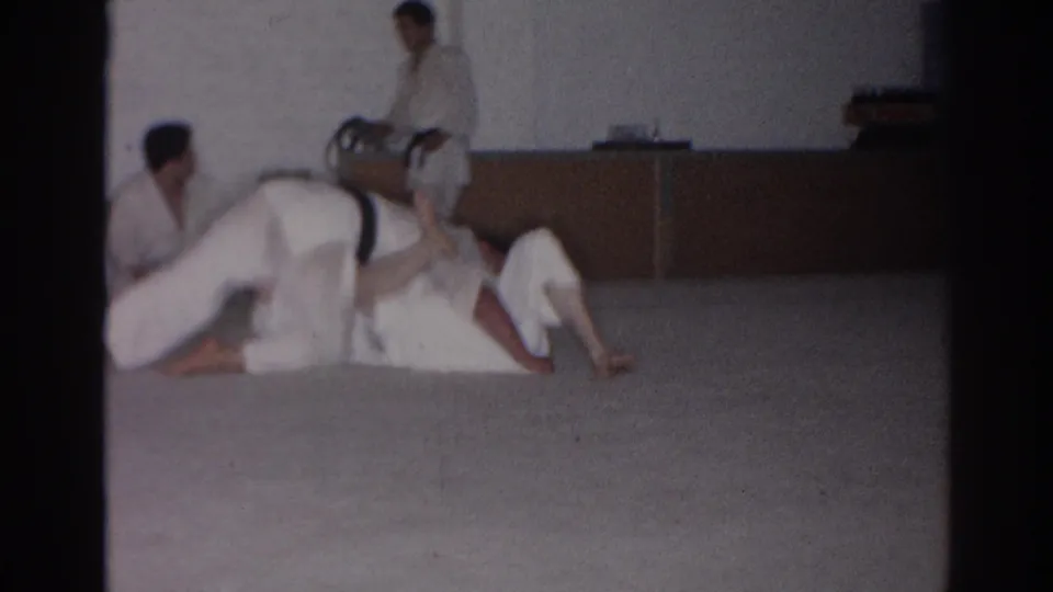During the San Gabriel martial arts practice, one person in a white gi bridges over a partner on the mat while observers sit or stand nearby.