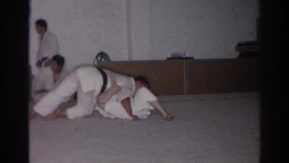 A white-gi grappler presses over a partner on the mat in the 1966 San Gabriel training room, with a seated observer partly visible.