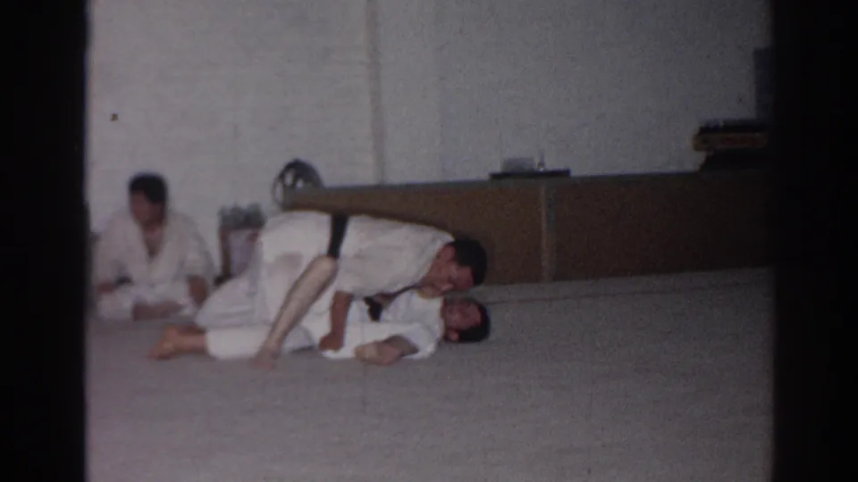 Near the end of the brief San Gabriel dojo clip, one martial artist appears to control a partner on the mat as a seated participant watches.