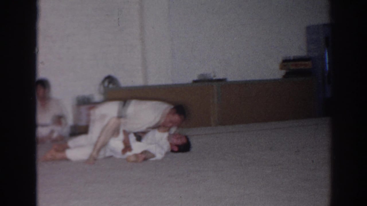 Two men in white gi uniforms grapple on an indoor mat while another person sits in the background.