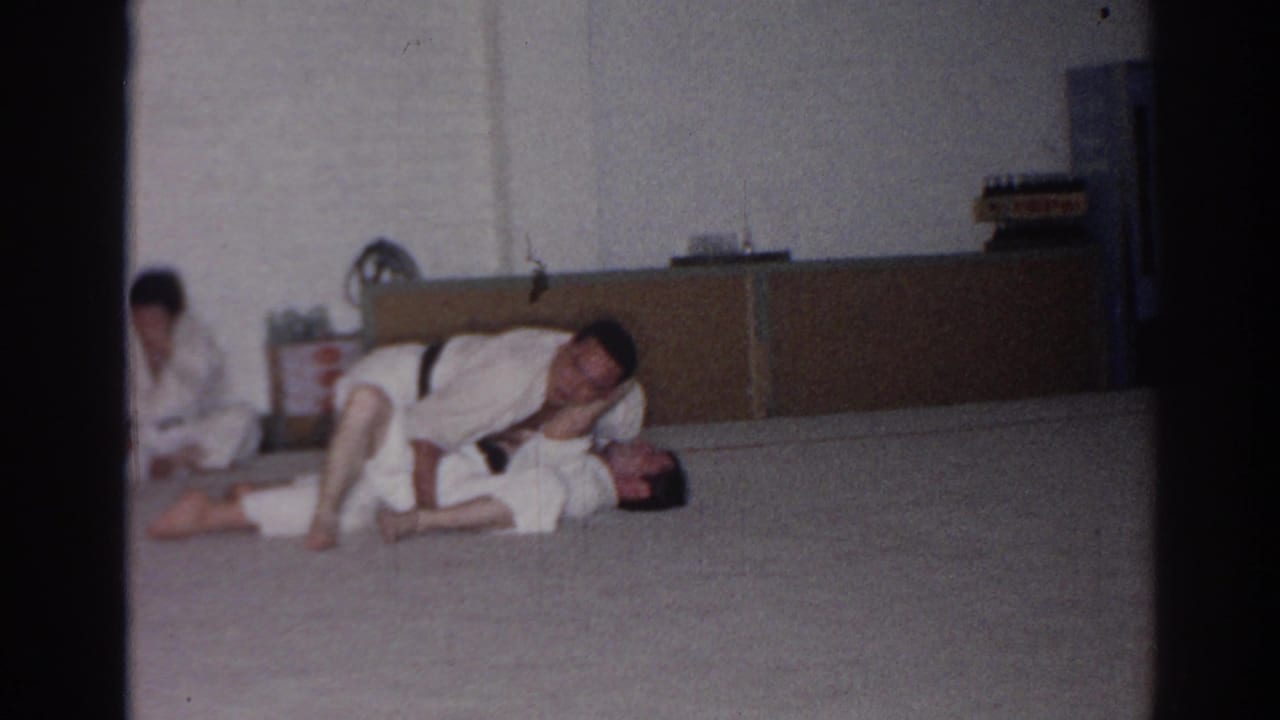 A man in a white gi leans over another person on a mat during a ground practice hold.