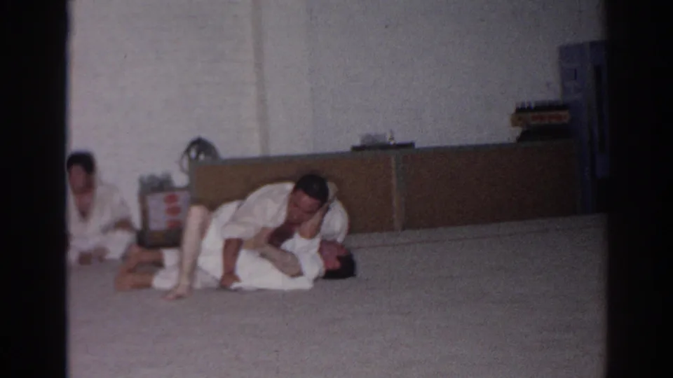 1966 San Gabriel home-movie frame showing a martial arts ground technique in a plain indoor training room.