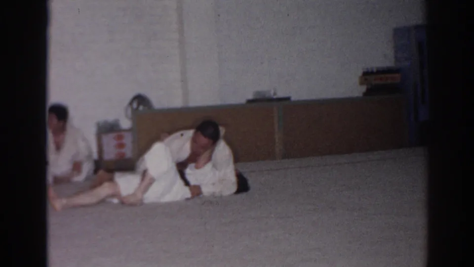 1966 San Gabriel frame of two gi-clad adults finishing a ground technique in an indoor practice space.
