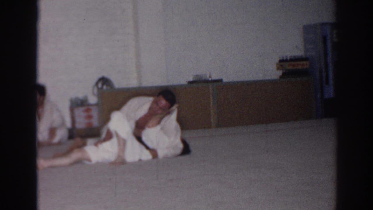 In San Gabriel, California, 1966 footage, two adult men in white gis grapple near the floor while a partial gi-clad figure appears at left.