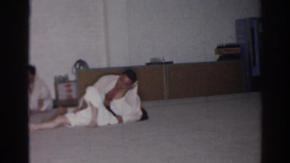 In San Gabriel, California, 1966 footage, two adult men in white gis grapple near the floor while a partial gi-clad figure appears at left.