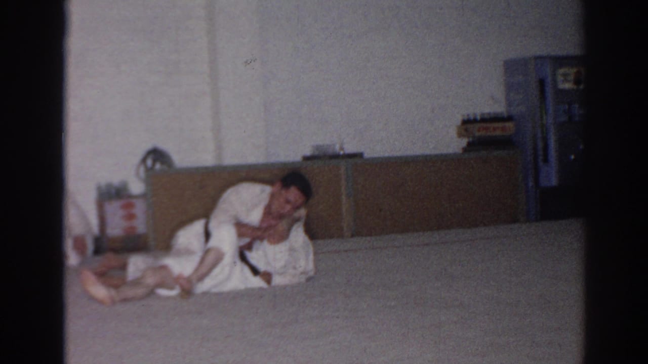 Two adult men in white gis continue grappling on a padded floor in the San Gabriel training room, with cabinetry and a blue upright fixture behind them.