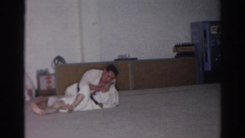 Two adult men in white gis continue grappling on a padded floor in the San Gabriel training room, with cabinetry and a blue upright fixture behind them.