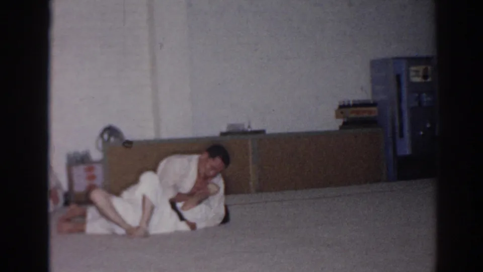 The 1966 San Gabriel home-movie frame shows one adult man in a white gi controlling another during a ground grappling drill.