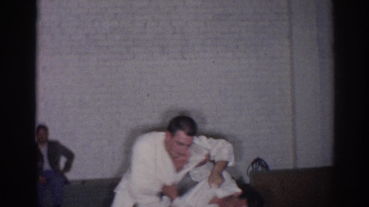 Two men in white judo uniforms grapple on an indoor mat while others sit nearby.