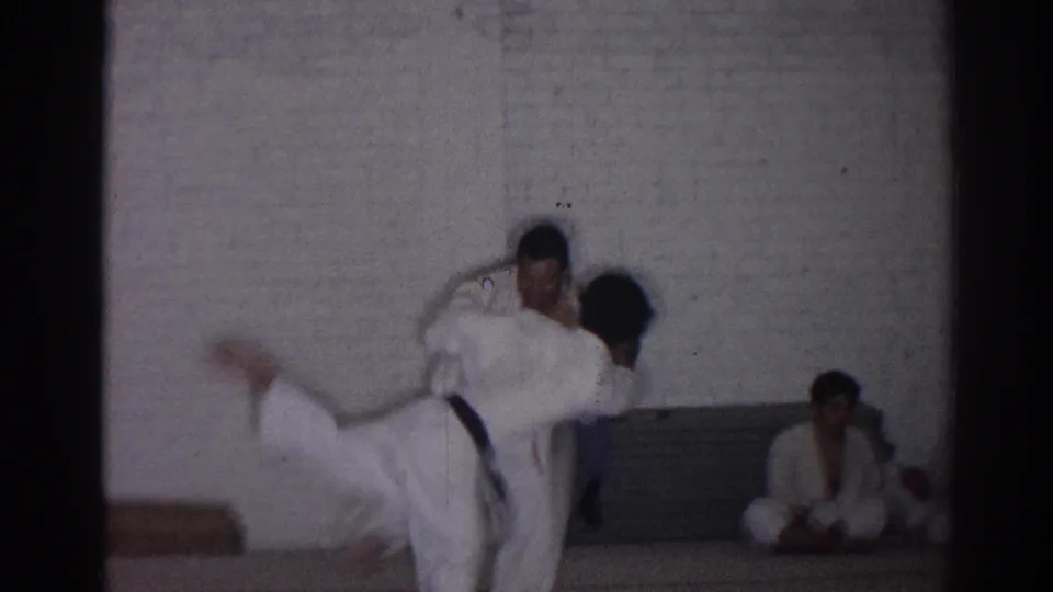 1966 San Gabriel dojo frame showing one judoka lifted or thrown while other practitioners sit along the wall observing.