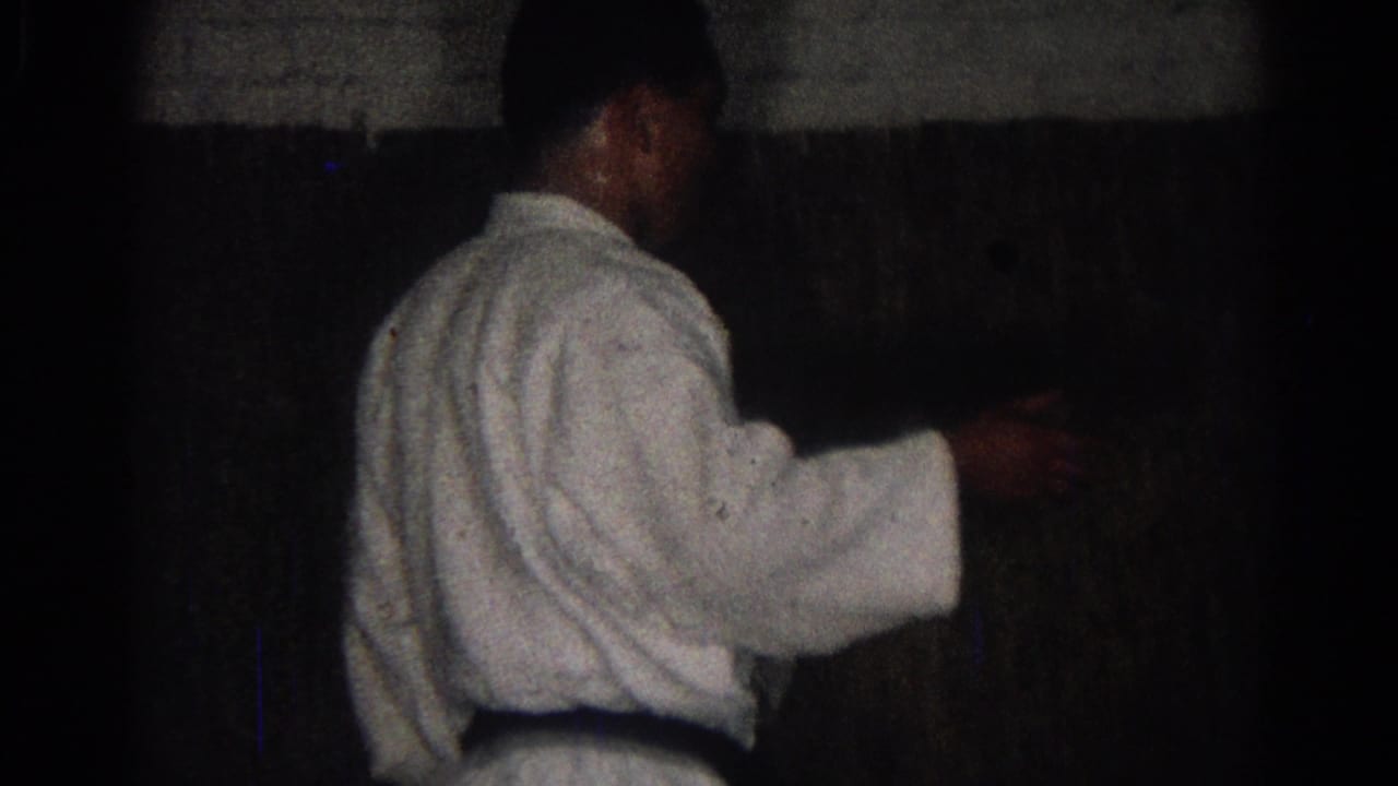 Man in a white gi and dark belt stands in a dim dojo.