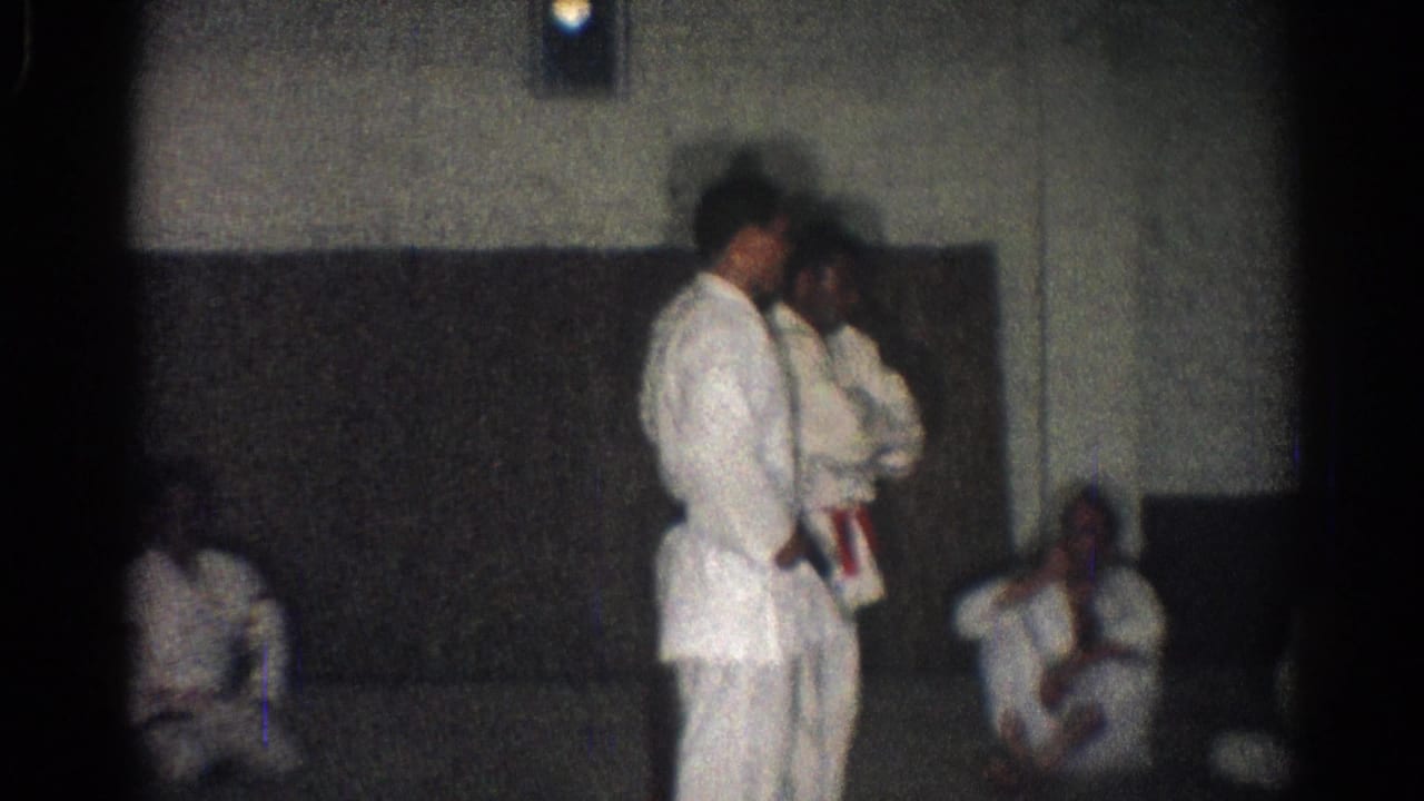 Two men in gi stand while others sit along the dojo wall.