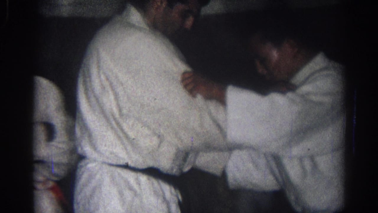 Two men in white martial arts uniforms grip each other in a dim indoor practice space.