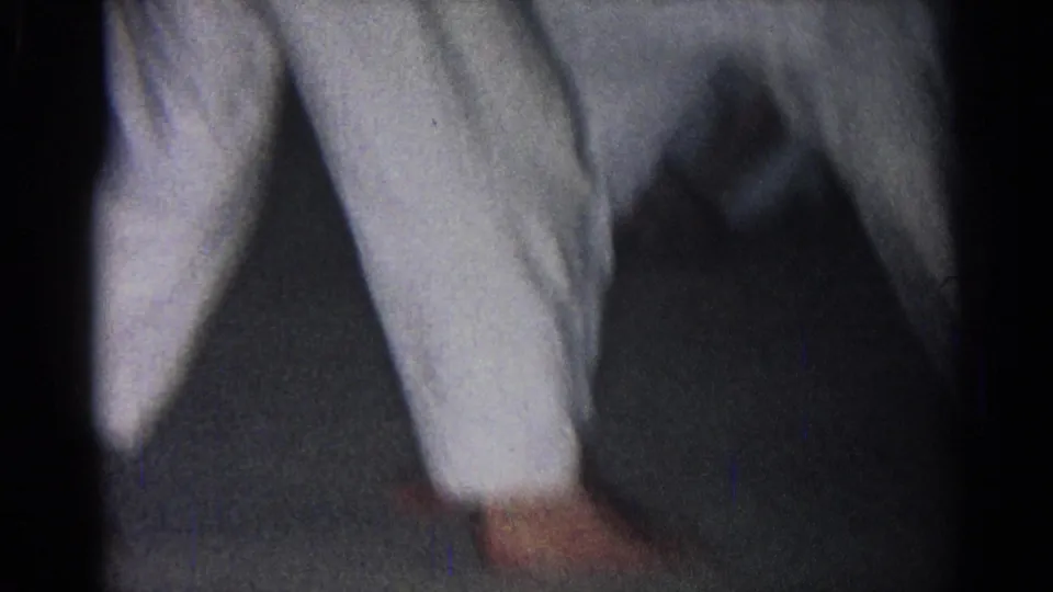 Close 1966 home-movie detail of martial arts footwork, with white gi pants and bare feet on a dark indoor floor.