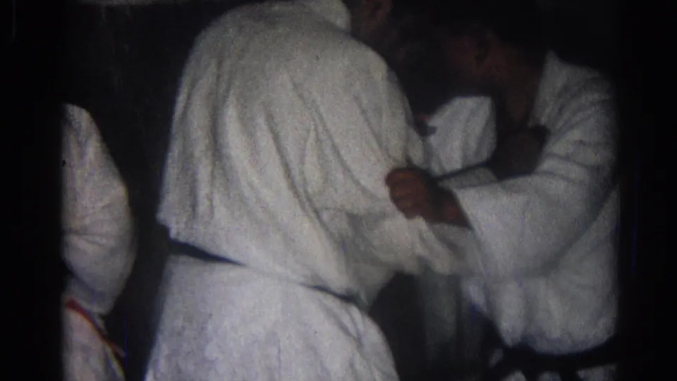 1966 San Gabriel home-movie frame showing two adult men in gi and black belts holding a close practice grip indoors.