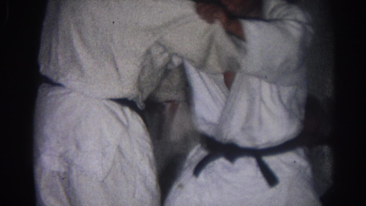 In 1966 San Gabriel footage, two adult karate practitioners in white gi and black belts grapple at close range indoors.