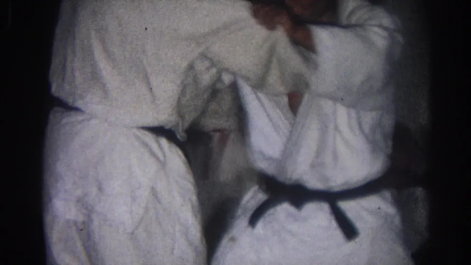 In 1966 San Gabriel footage, two adult karate practitioners in white gi and black belts grapple at close range indoors.