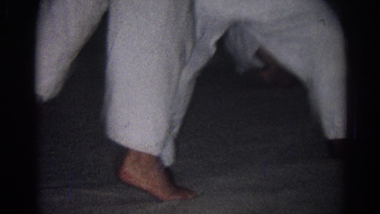 A low view shows a bare foot and white gi pant legs on a dark indoor floor during the San Gabriel karate practice.