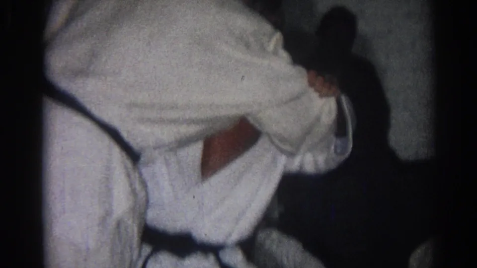 Two adult men in white gi continue a close grappling drill, with bodies bent forward and partially obscured by the handheld framing.