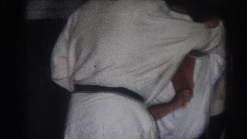 A practitioner in a white gi leans over another adult during a close martial arts hold in the 1966 indoor training footage.