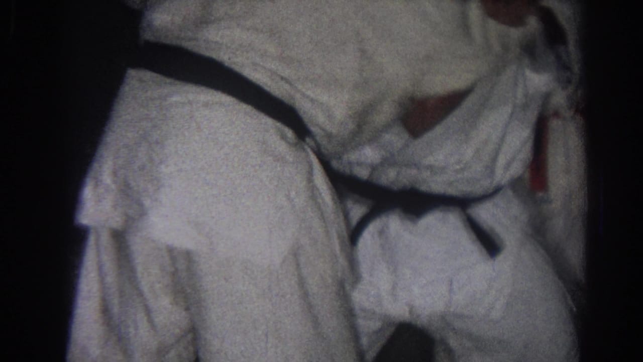 Close 1966 home-movie frame of white gi uniforms and black belts during a martial arts grappling moment indoors.