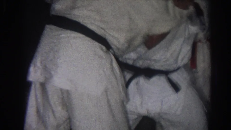 Close 1966 home-movie frame of white gi uniforms and black belts during a martial arts grappling moment indoors.