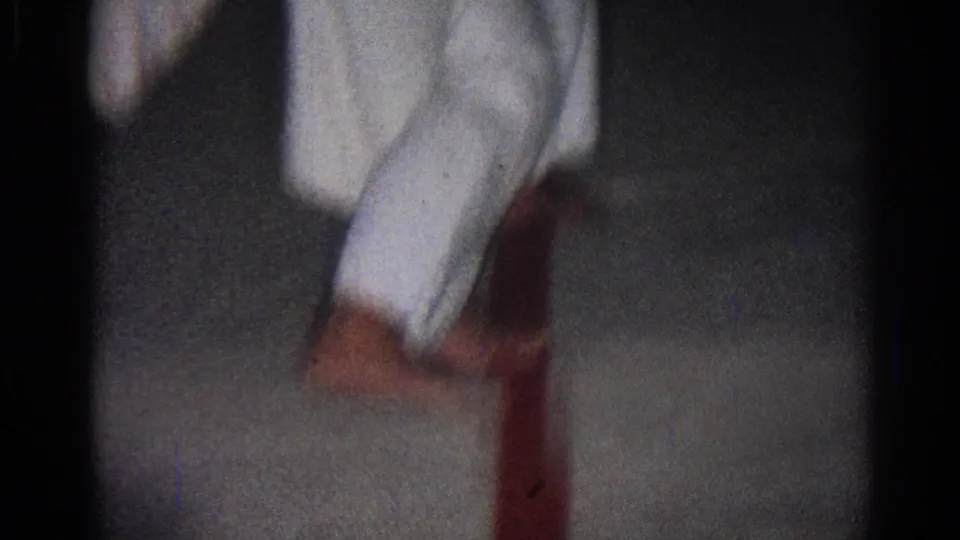 A martial artist in a white gi crouches low beside another partly visible person during the dim indoor San Gabriel practice footage.
