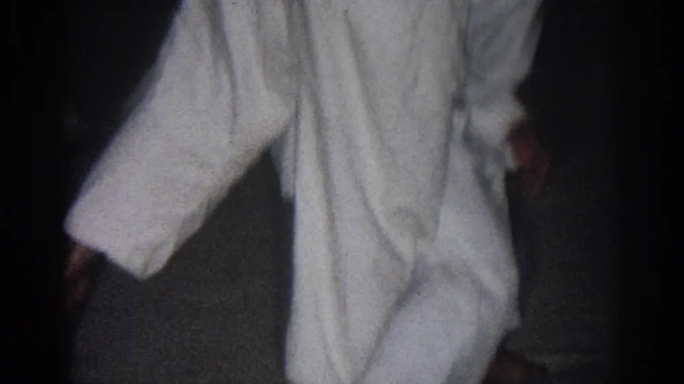 A white gi fills the frame as the practitioner moves indoors, with the dark floor and soft 8mm grain visible.