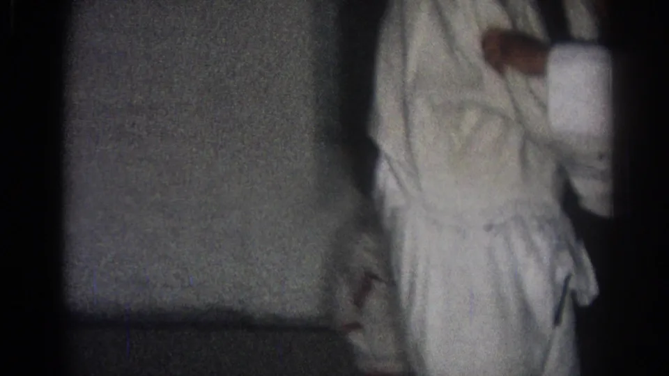 A judo practitioner in a white gi appears at the edge of the dim indoor frame, with the training space mostly dark.