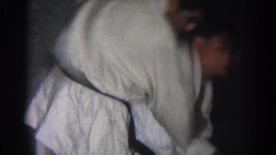 A judo practitioner in a white gi bends forward during a technique, with another adult partly visible at the side of the dim room.