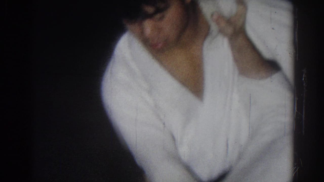 A man in a white martial arts uniform bends forward while grappling with another person.