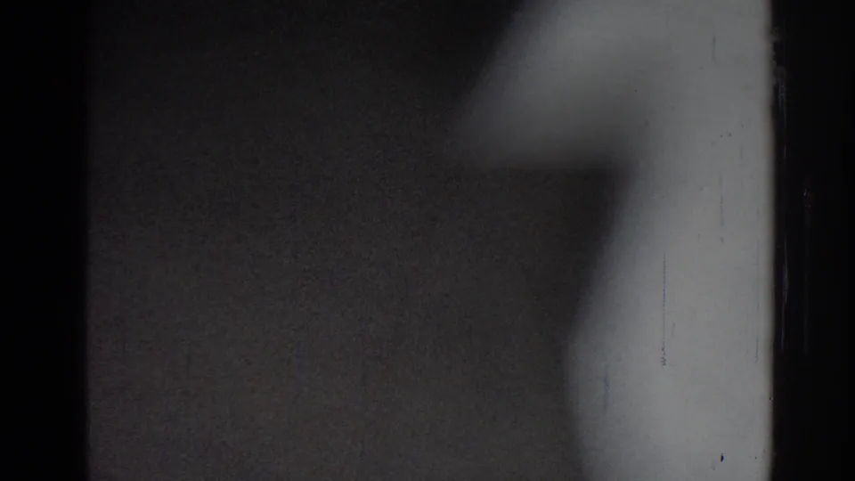 Soft 8mm frame from the San Gabriel clip showing a white-uniformed practitioner moving through shadowed indoor space.