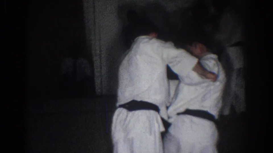 Two boys in white martial arts uniforms and dark belts are seen from behind in a dim San Gabriel indoor practice setting.