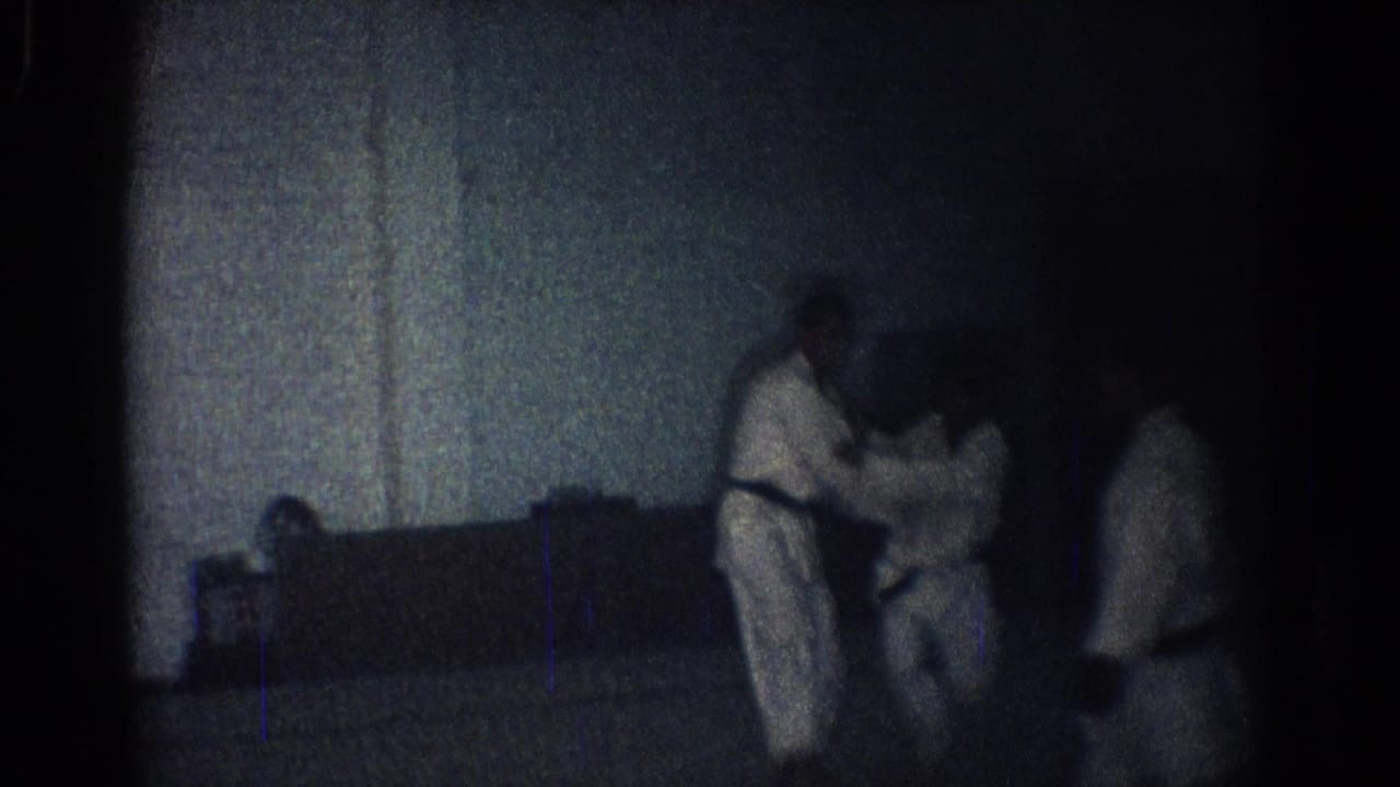 Dim dojo scene with two men in karate uniforms practicing a hold near mats.